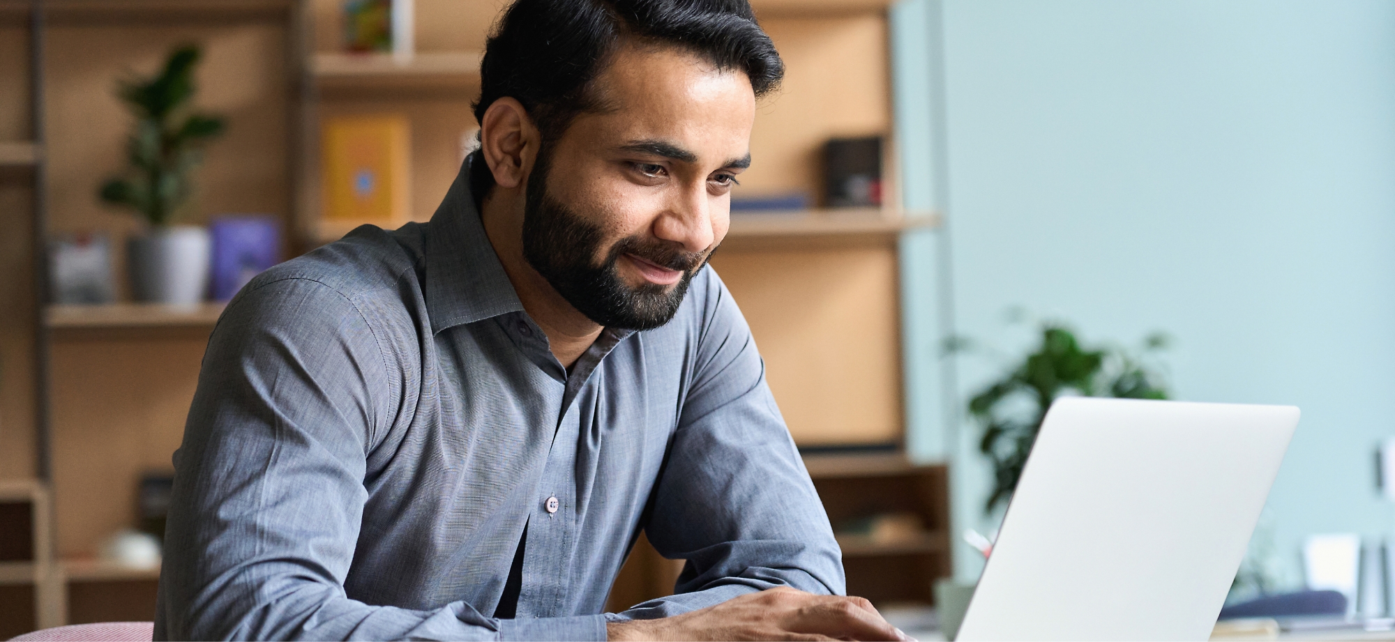 A man with a beard smiling lightly while working on a laptop in an office with shelves in the background.