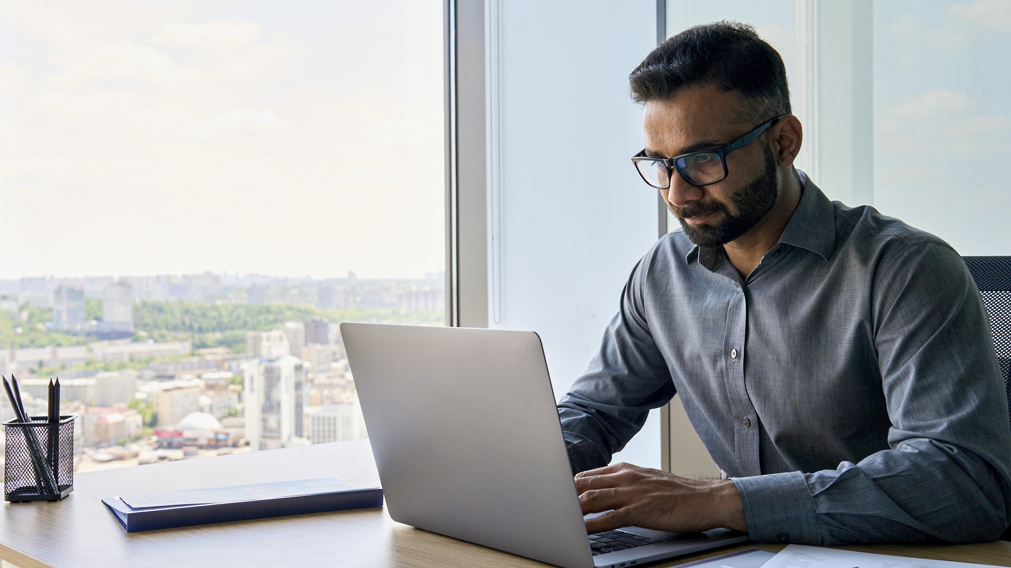 Egy szemüveges férfi elmélyülten dolgozik egy laptopon egy ablak melletti íróasztalnál, ahonnan kilátás nyílik a városra.