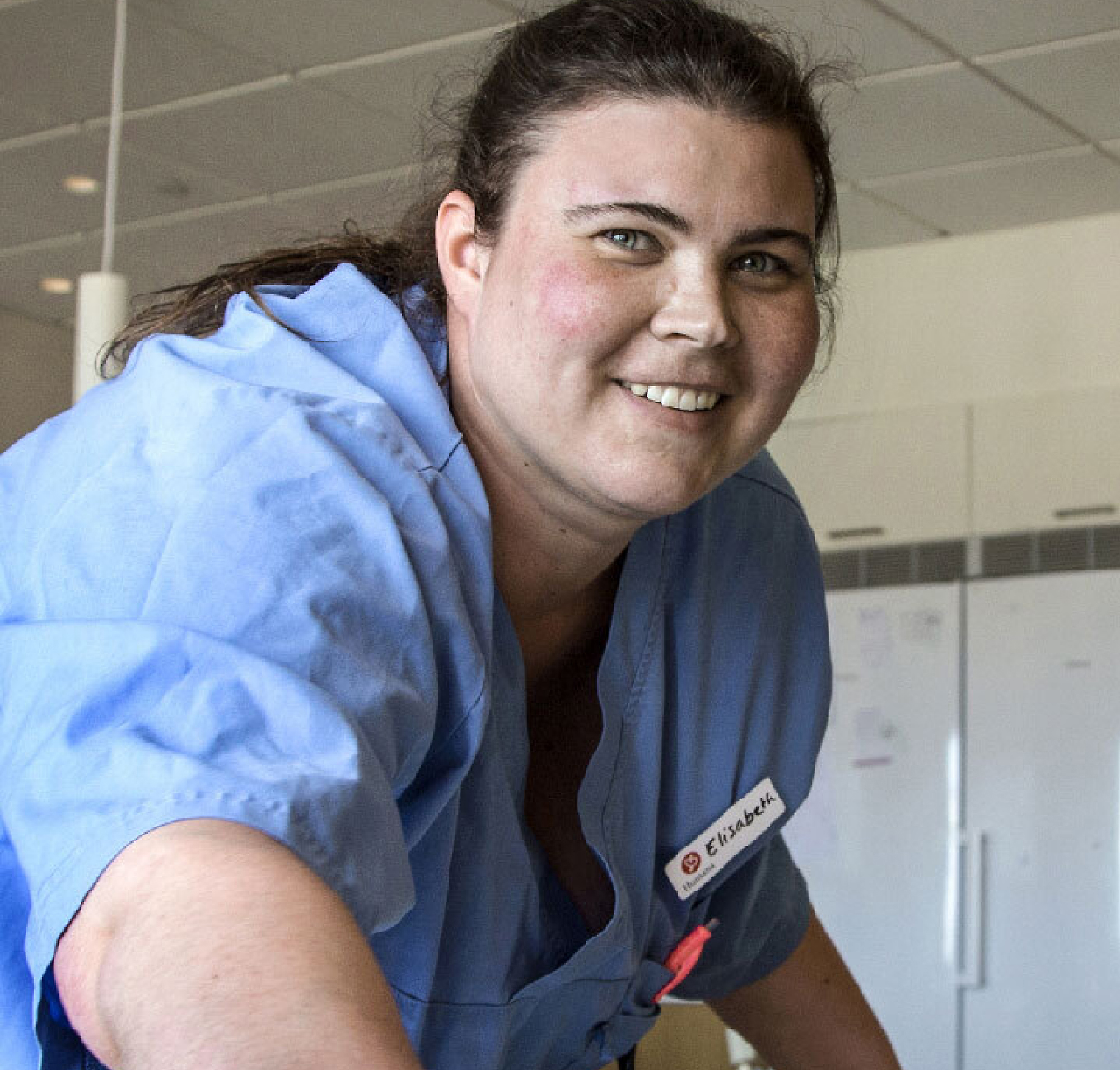 Een verpleegster met een naamplaatje met de tekst 'Elizabeth', glimlachend in een blauw uniform, in een lichte, ruime ziekenhuisomgeving.