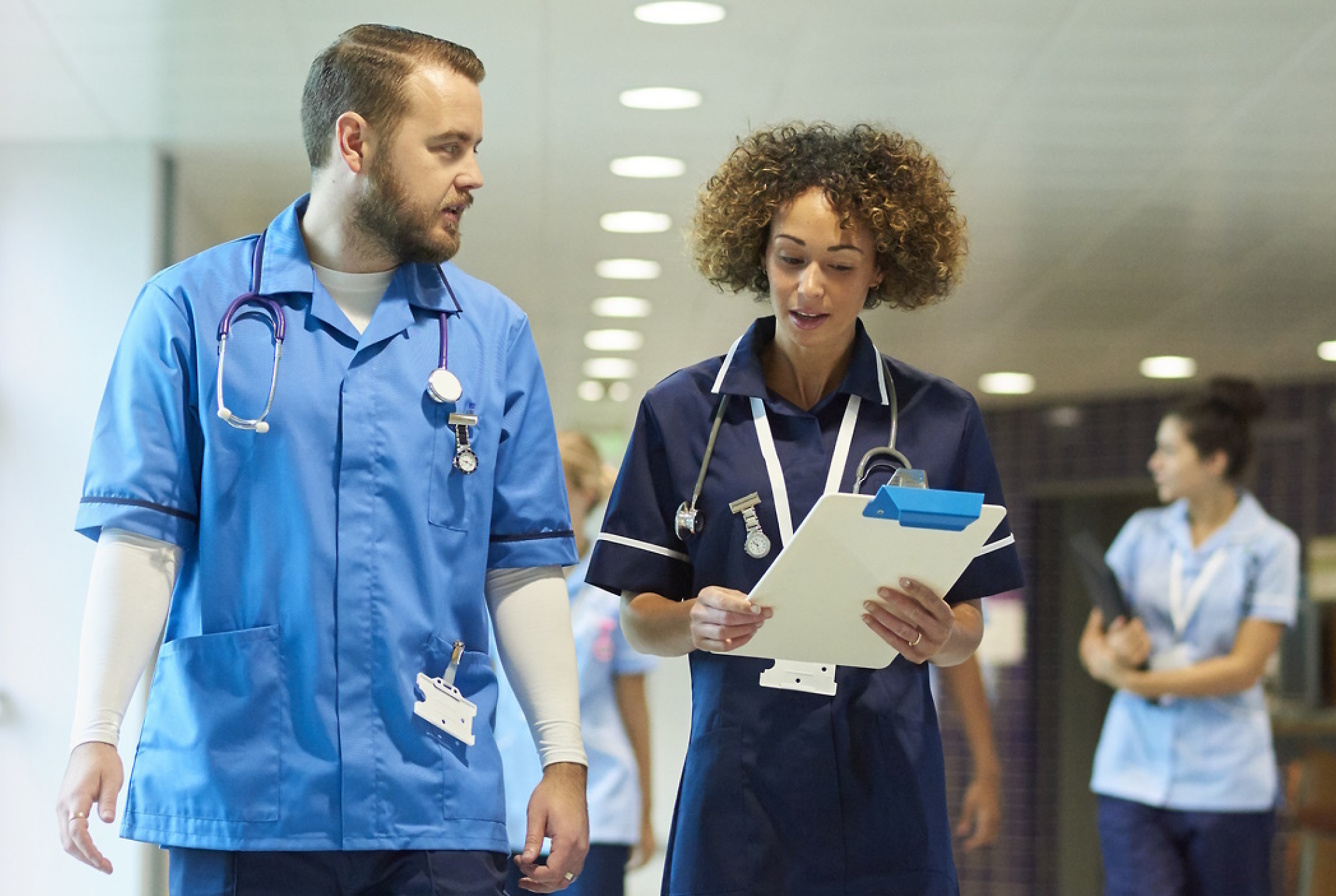 Двама медицински специалисти в сини униформи разговарят и вървят в болничен коридор, като единият държи клипборд. 