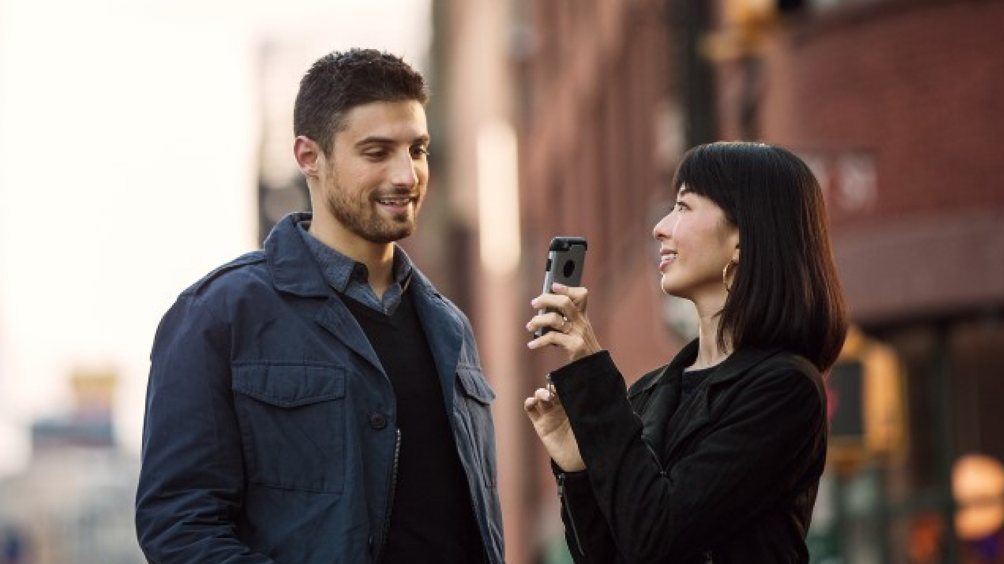 Женщина, держащая смартфон, направляет его на мужчину, который оба стоят на городской улице, улыбаются и разговаривают.