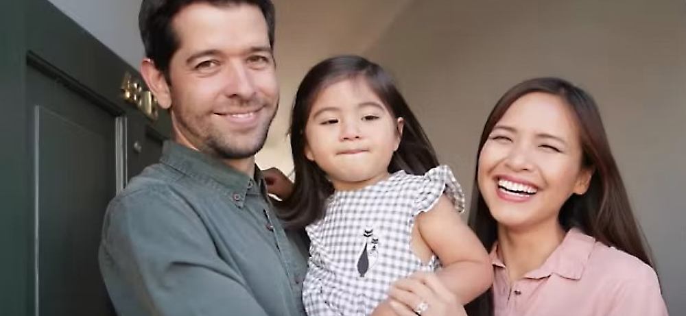 Een gelukkig gezin met een man, vrouw en jong kind glimlachend naar de camera, staande bij de ingang van een huis.