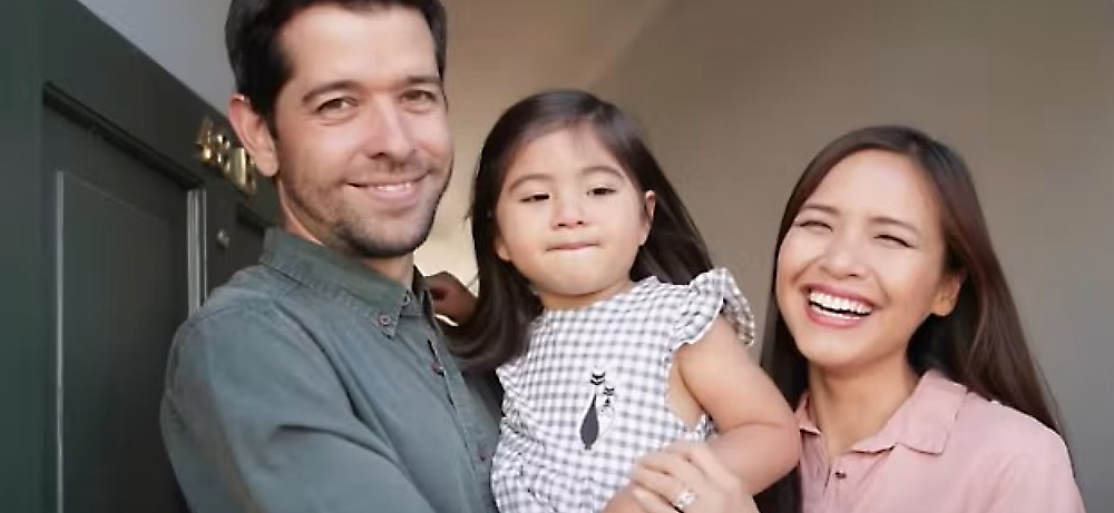 A happy family with a man, woman, and young child smiling at the camera, standing at a home entrance.