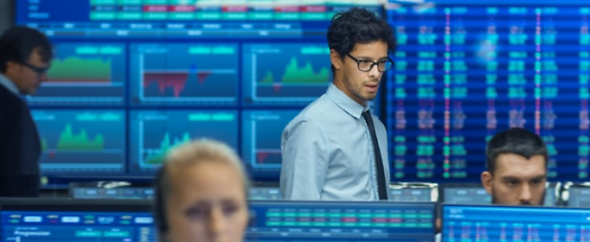 Man in glasses and a tie standing in a busy stock exchange office with financial data on multiple screens in the background.