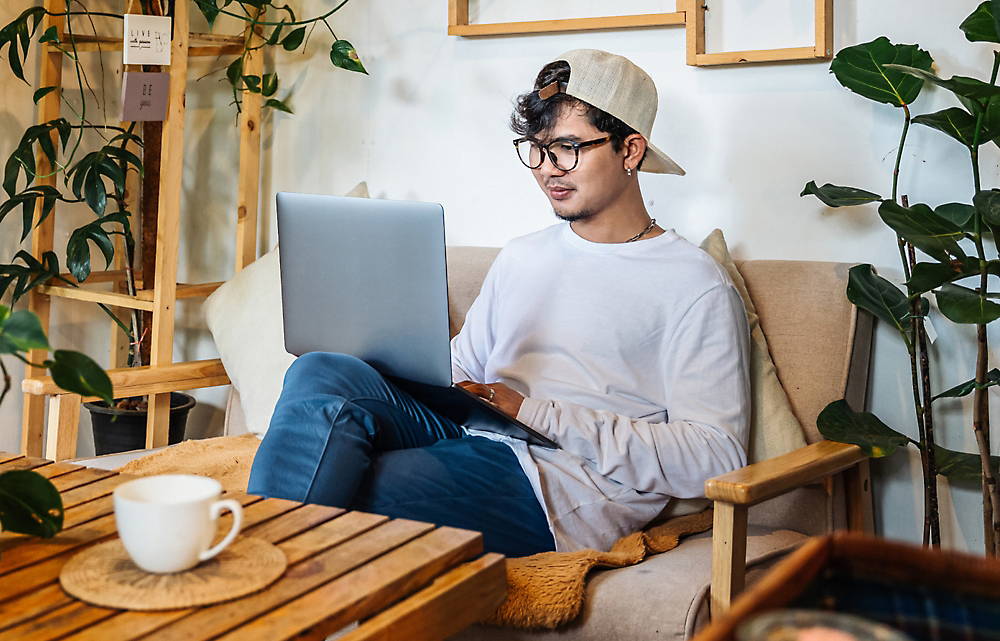 A person uses a laptop while sitting in a room with plants and wooden furniture.