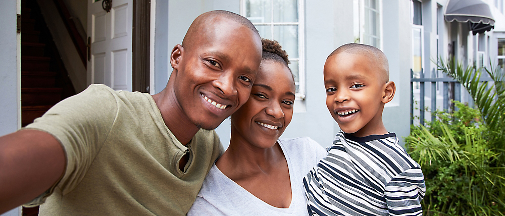 A smiling family—a man, woman, and boy—poses for a selfie, standing in front of their home.