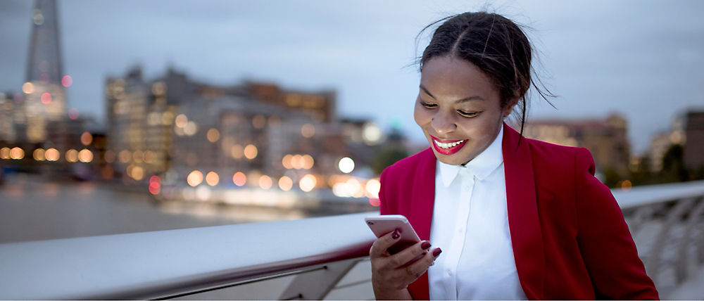 たそがれ時に市街地の橋の上で笑顔でスマートフォンを使っている赤いブレザー姿の若い女性。