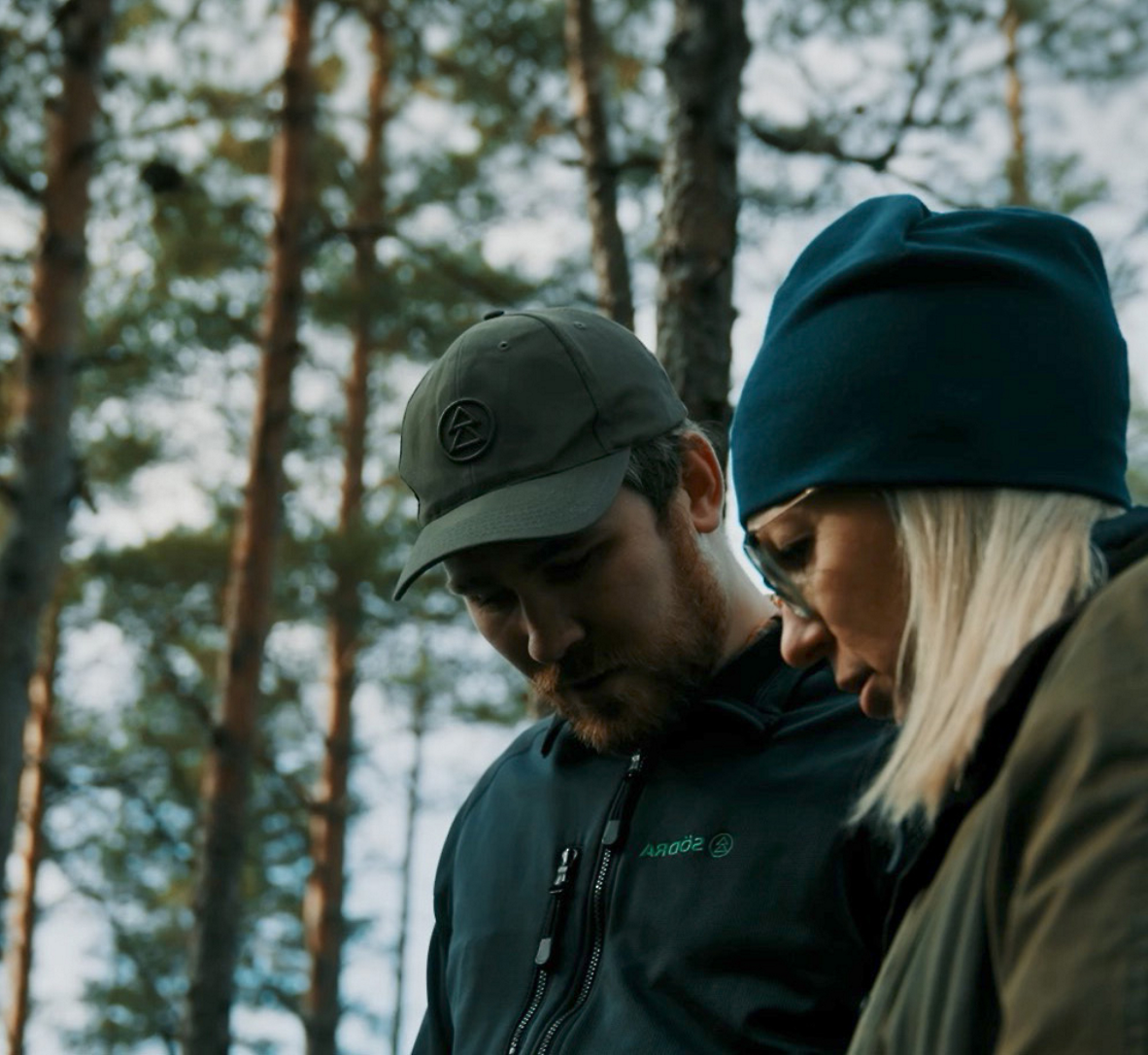 Two people wearing outdoor clothing stand closely together in a forest, looking down at something.