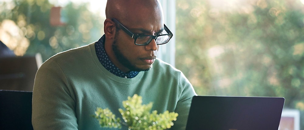 A man wearing glasses and a green sweater is focused on working on a laptop at a table, with a potted plant in the foreground.