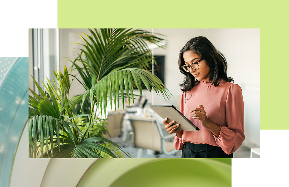 Uma mulher usando óculos e uma blusa rosa lendo um tablet em um escritório moderno com grandes janelas e plantas.