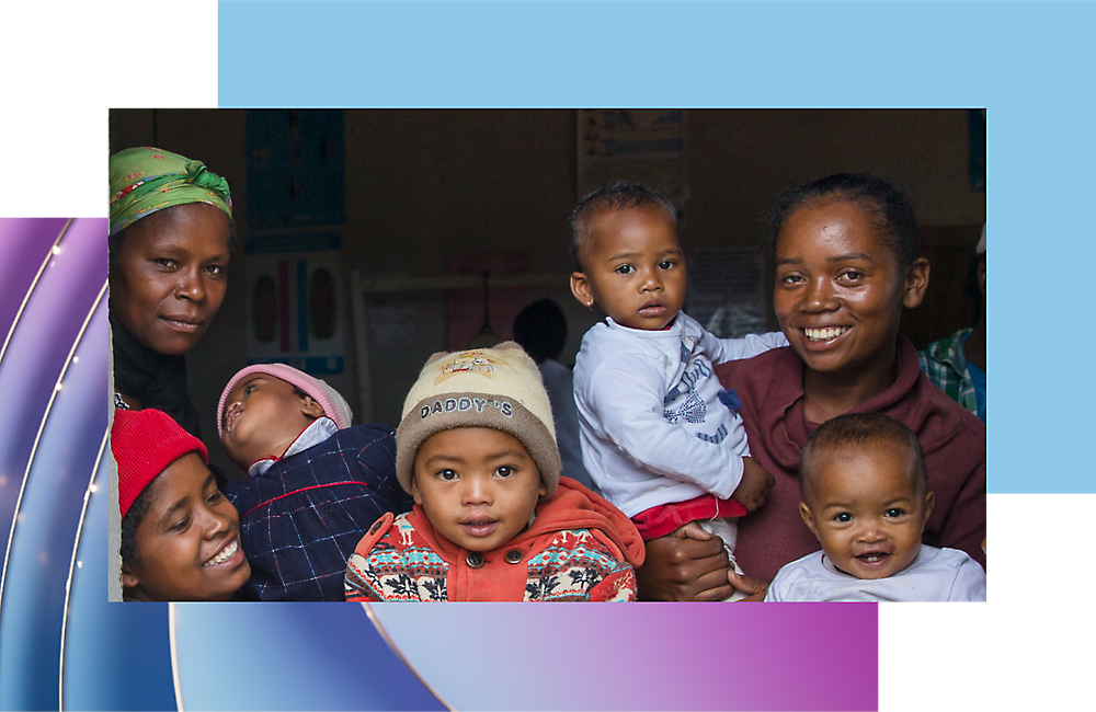 Three women and four children in a room, smiling and looking towards the camera; one child is on a woman's back