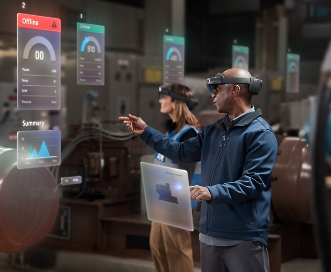 To personer, der bruger augmented reality-headsets til at interagere med virtuelle dataskærme i et industrielt miljø.