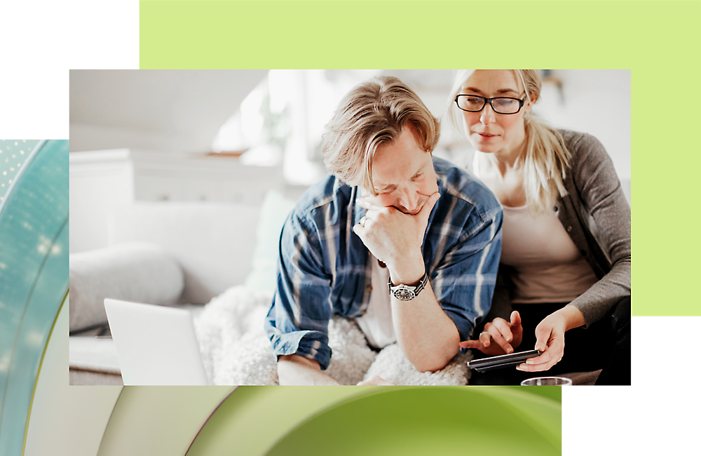 A middle-aged couple reviews documents on a digital tablet, seated on a sofa in a bright, cozy living room.