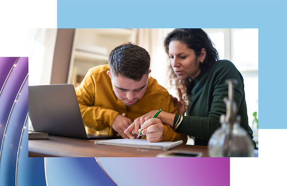 Two people, a young man and a woman, collaborate over documents with a laptop on a wooden table in a brightly lit room.