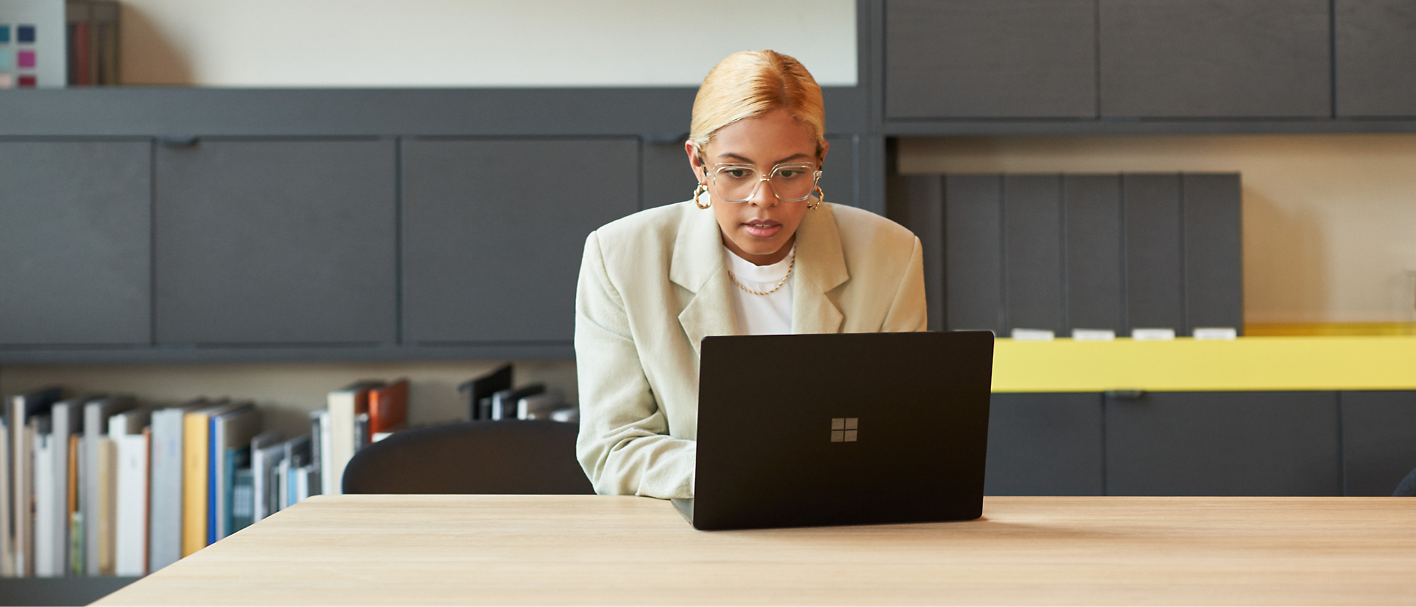 A person with short blonde hair and glasses sits at a wooden table, focused on a black laptop