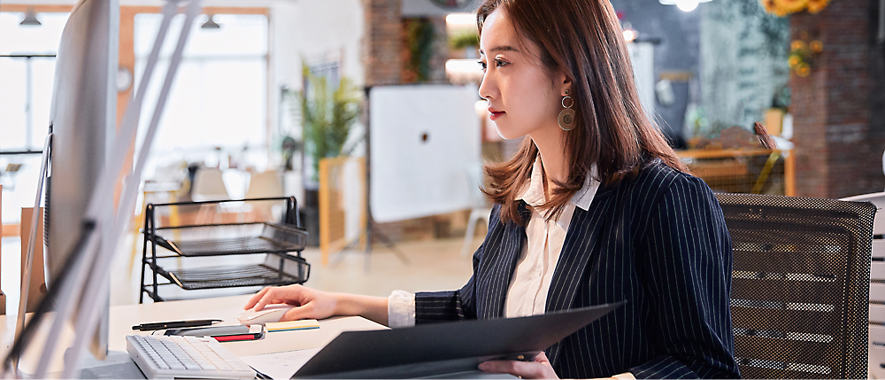 A woman in a business suit works at a desk, looking at a computer screen and holding a folder. 