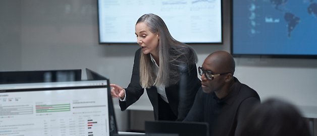 A woman presenting in a meeting with a man and digital screens displaying data in the background.