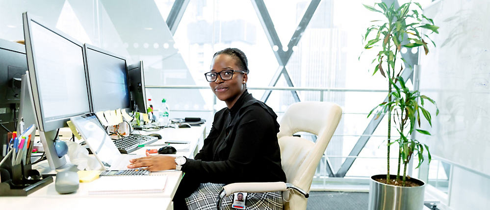 Vrouw met bril die aan een bureau werkt met meerdere beeldschermen in een modern kantoor met geometrische vensters en een potplant.