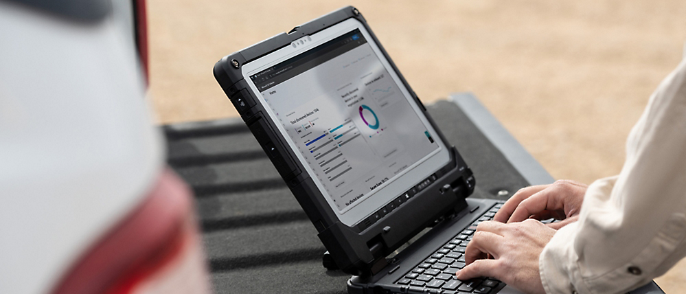 A person works on a rugged laptop displaying graphs, sitting outdoors on a sandy surface.