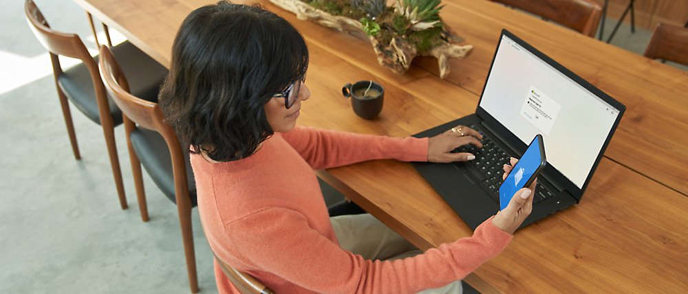 Een vrouw in een oranje trui die een smartphone en laptop gebruikt aan een houten tafel.