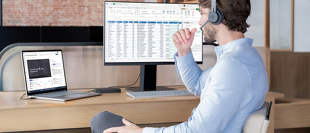 A man wearing a headset sits at a desk, working on a computer with spreadsheet data on the screen in a modern office 