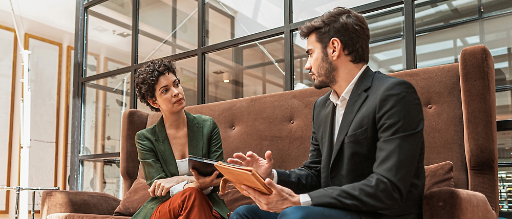 Two professionals discussing over a digital tablet while sitting on a couch in a modern office lobby.