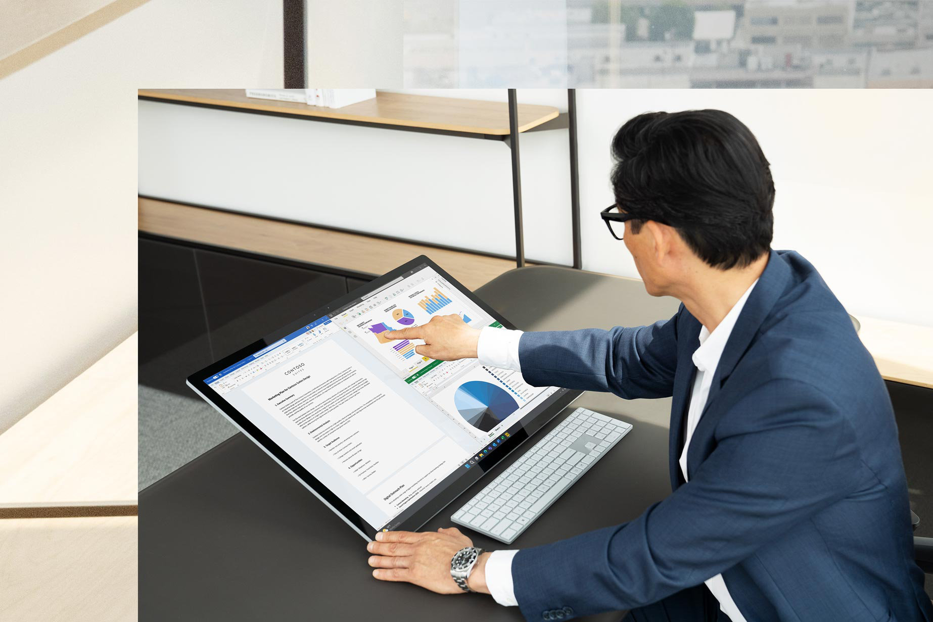 Työpöydän ääressä istuva mies koskettaa Surface Studio 2+:n näyttöä ja pitää yritysesitystä näyttö kallistettuna taakse studiotilaan.