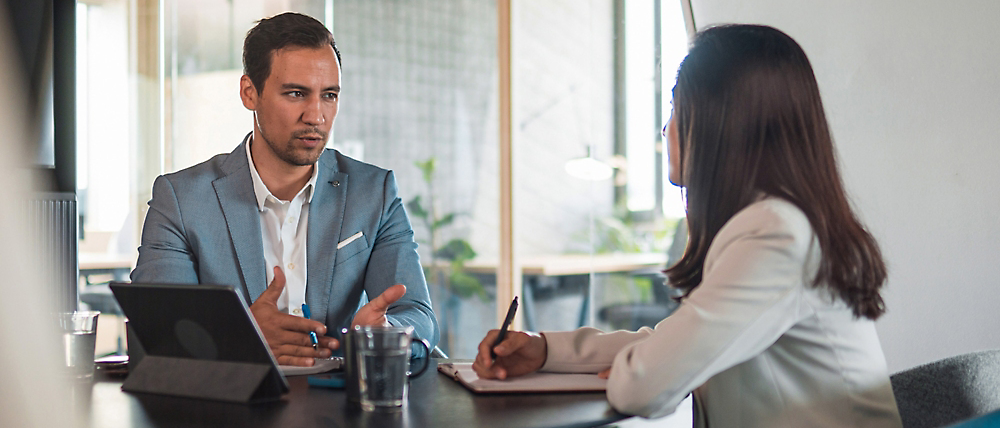 Two professionals in a meeting, a man speaking and gesturing while a woman listens and takes notes.