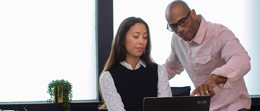 Two professionals, a man and a woman, analyze data on a laptop in a modern office setting.