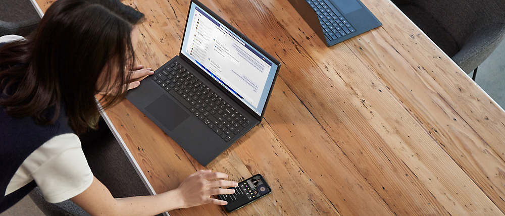 Een bovenaanzicht van een vrouw die op een laptop werkt aan een houten tafel met behulp van een draagbaar apparaat.