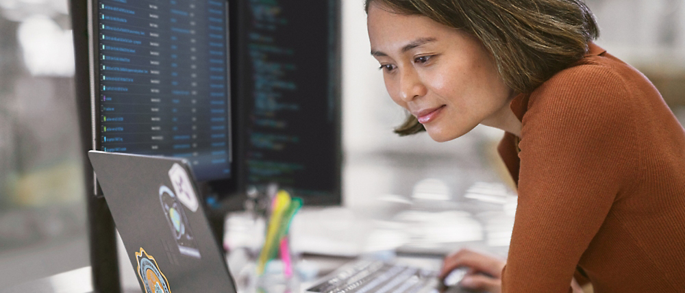 A woman closely examines coding on a computer screen in an office setting.