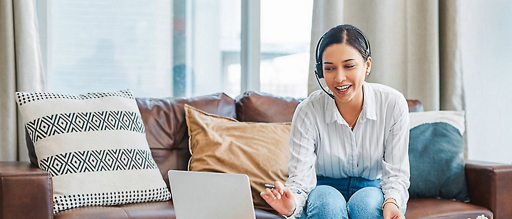 A cheerful woman sits on a sofa with a laptop, engaging in a conversation in a bright, modern living room.