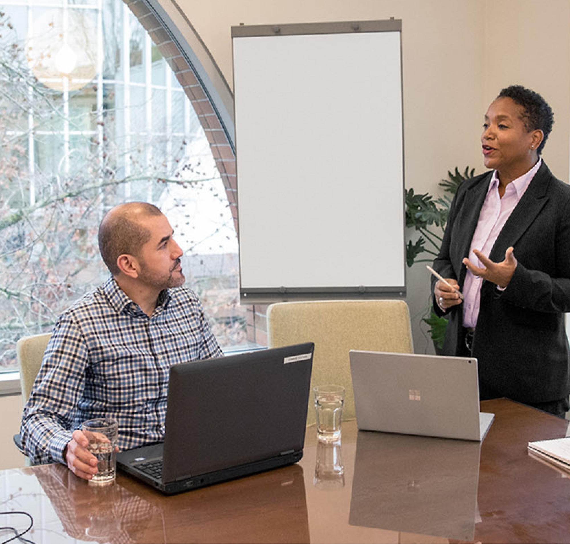 صورة لمتخصصين في غرفة الاجتماعات، أحدهما يقف لتقديم العرض والآخر يجلس على طاولة مع أجهزة كمبيوتر محمولة أمام السبورة البيضاء.