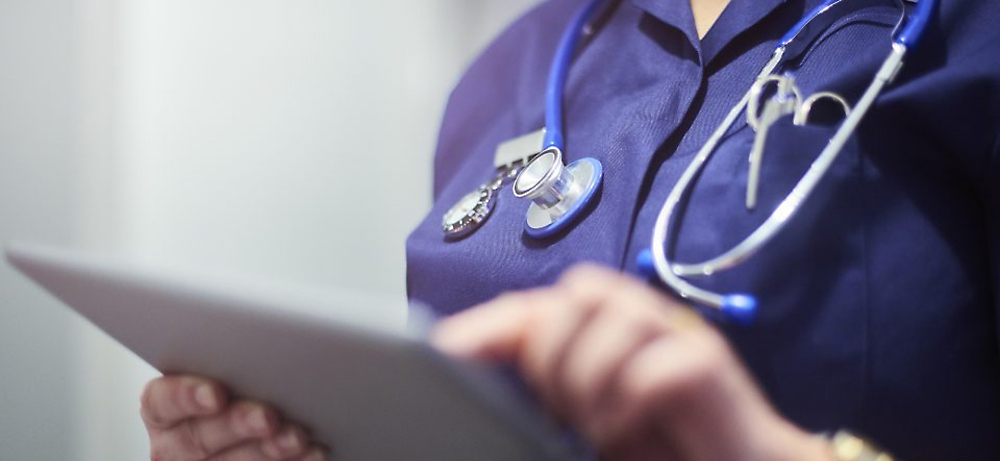 Doctor in a blue medical uniform holding a tablet, with a stethoscope hanging around the neck, reviewing medical records.