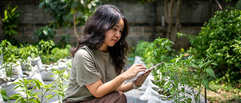 A woman crouches in a garden, holding a tablet and examining plants, surrounded by greenery.