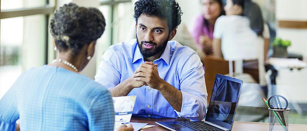 Two professionals engaged in a serious discussion at a table with laptops in a busy office setting.