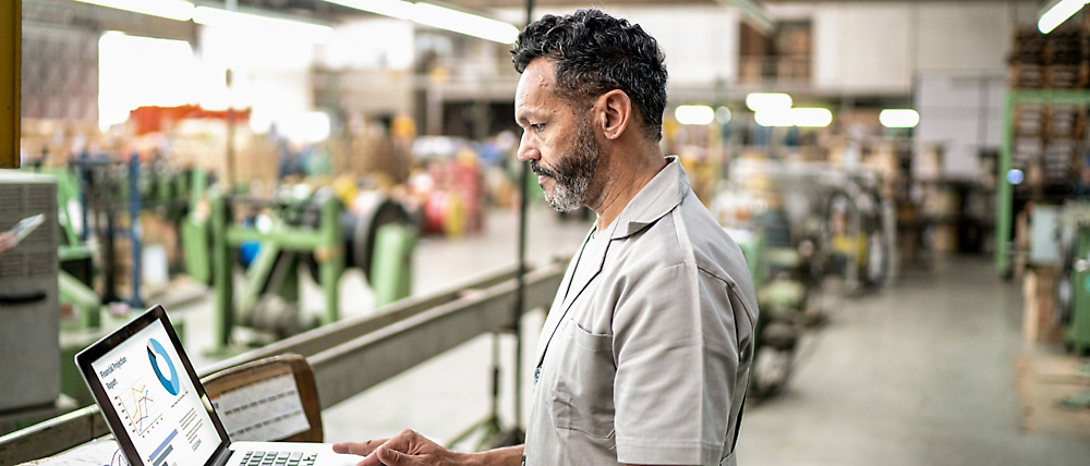 A middle-aged man with gray hair and beard, wearing a light gray shirt, reviews data on a laptop in an industrial warehouse.