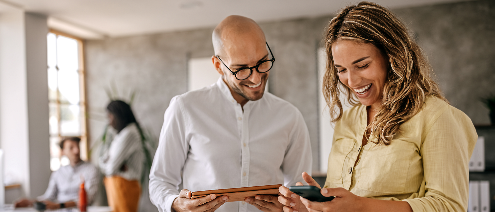 Duas pessoas em pé e sorrindo dentro de casa, segurando um tablet e um smartphone.