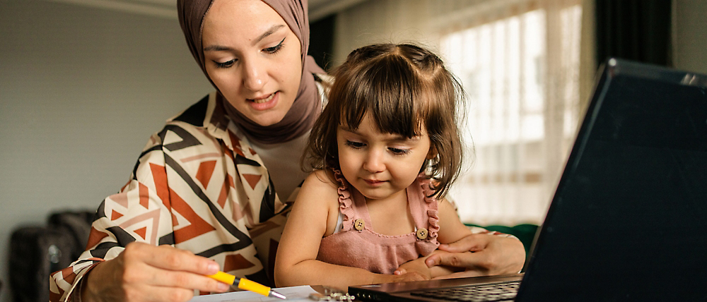Een vrouw met een hijab en een jong kind die aan een bureau zitten en samen naar een laptopscherm kijken.