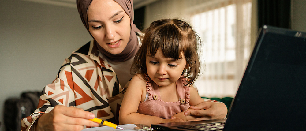 Femme portant un casque et un jeune enfant assis à un bureau, regardant un écran d’ordinateur portable ensemble.