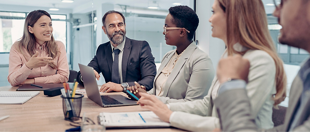 Diverse group of business professionals engaged in a meeting at a conference table with laptops and documents.