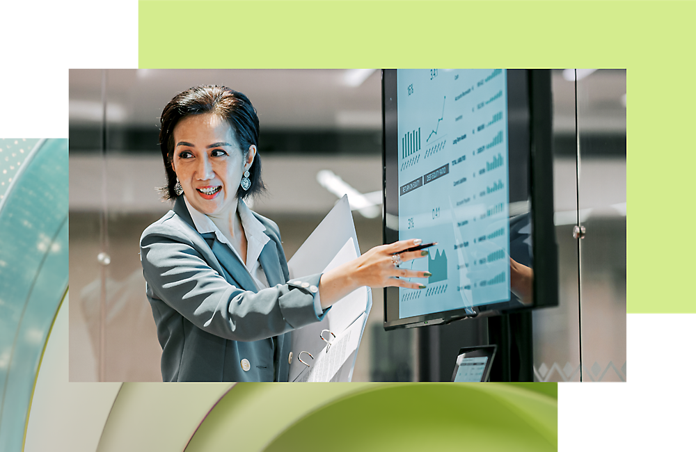 A professional woman presenting data on a digital screen, smiling and pointing, with a modern office background.