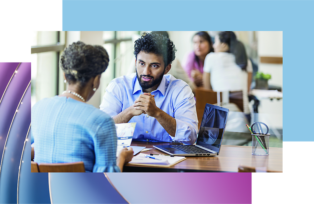 Two professionals engaged in a discussion at a table with laptops and documents in a brightly lit office setting.