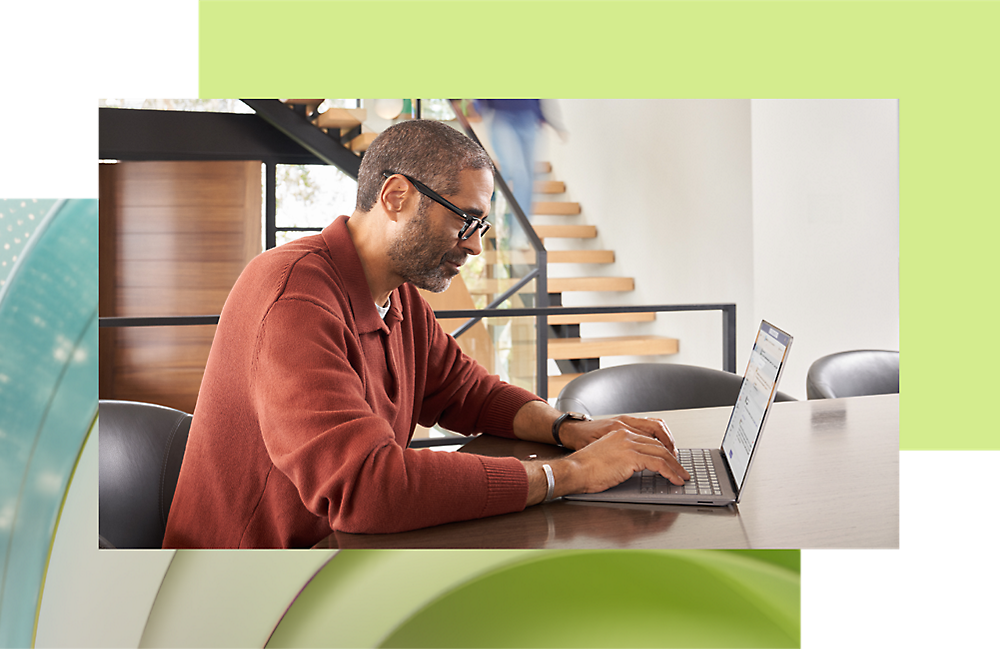 Een man met een bril en een rood vest werkt op een laptop aan een modern bureau in een helder verlichte kantoorruimte.