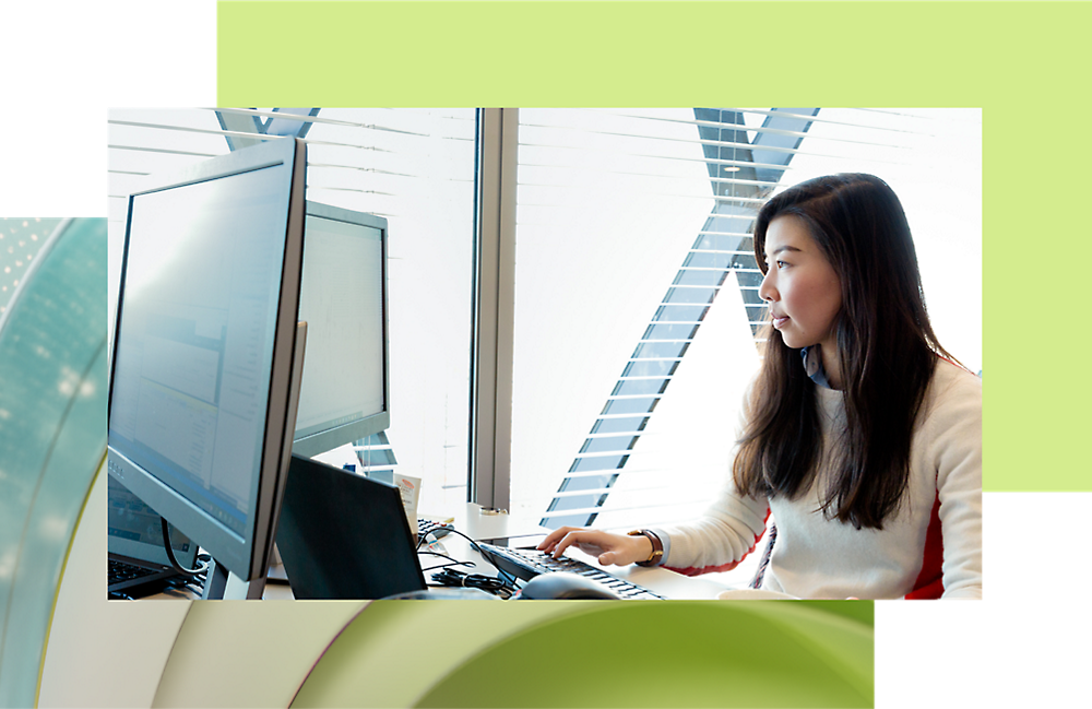 A woman with curly hair working at a desk, using a computer in a brightly lit office environment.