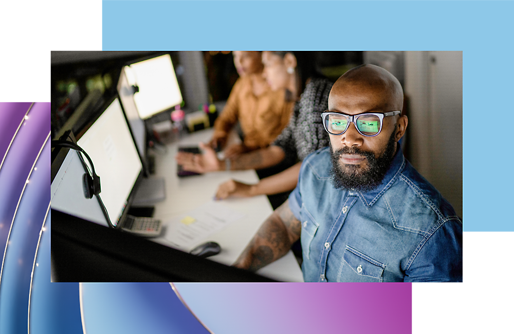 A man with a beard wearing glasses looks at the camera from a busy office setting with two colleagues