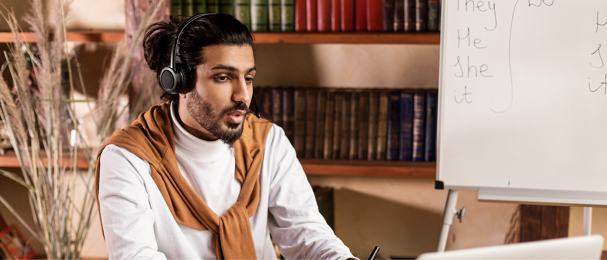 Uma pessoa com uma barba e cabelo longo, usando fones de ouvido, em uma mesa com um laptop. 