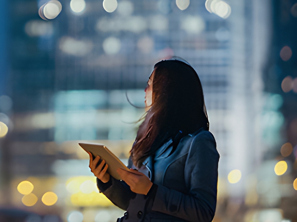 Une femme en costume d’affaires tenant une tablette regarde vers le haut dans la nuit, éclairée par les lumières de la ville