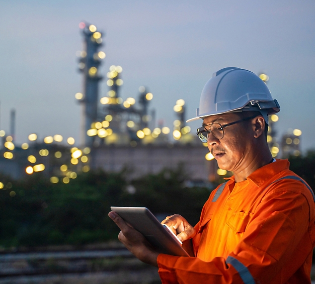 Ingeniero con un mono naranja y un casco que usa una tableta, con una planta industrial iluminada al atardecer en segundo plano.