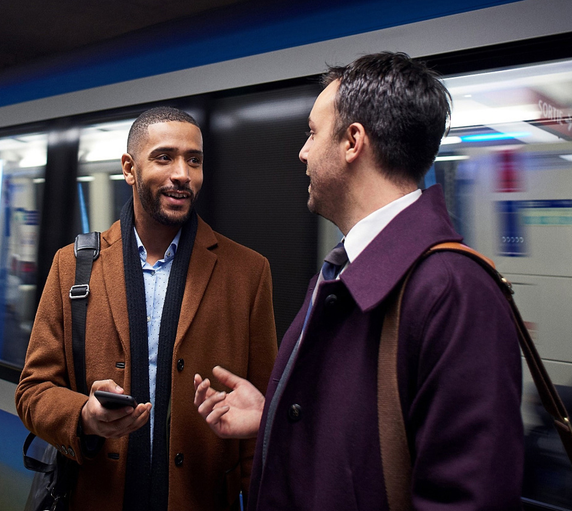 To menn som snakker på en undergrunnsplattform, med én som holder en smarttelefon og et tog uskarpt i bevegelse bak seg.